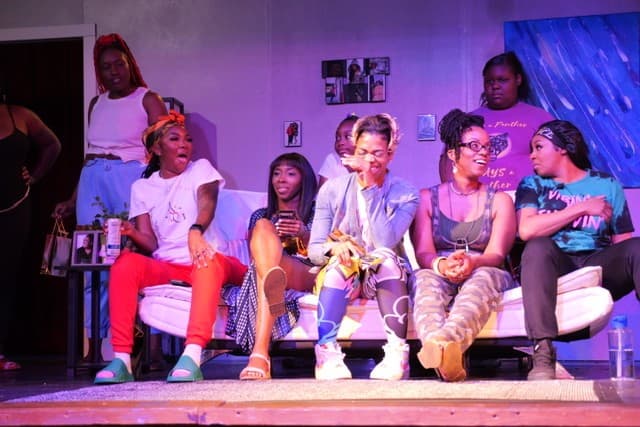 The Aunt Sarah’s Girls ensemble in performance, gathered around a sofa on the living-room set under stage light.